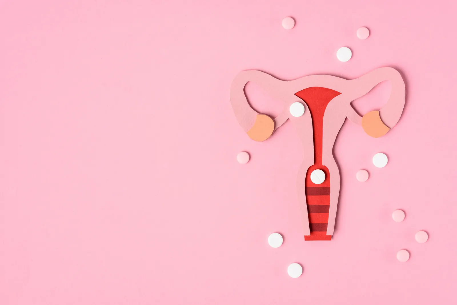 Paper cut-out of a uterus surrounded by pills on a pink background